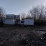 EIne schlammige Fläche. Fahrspuren von schwerem Gerät, Bagger oder Traktor. Der Boden ist feucht. Am Ende der Schlammfläche stehen verwahrlost wirkende, nicht mehr bewohnte Container, die von rechts von der Morgensonne beschienen werden. Dünne Leitungen einer Hochspannungstrasse am blassblauen Himmel. Rechts und links der Container ragen die Zweige von Bäumen in die frische Luft, noch unbelaubt und feingliedrig.