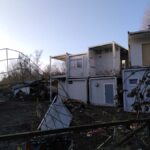 EIn verbogener, niedergedrückter Zaun aus einer Bewehrungsmatte im Vordergrund. Stachelig anmutendes Gestrüpp. Ausgediente Baucontainer oder Flüchtlingsheimcontainer im Zentrum des Bildes. Im Hintergrund ein Baumwqipfel in der frühen Morgensonne. Frische Frühlingsluft und Vogelzwitschern, ein Rotkehlchen wohlmöglich.  